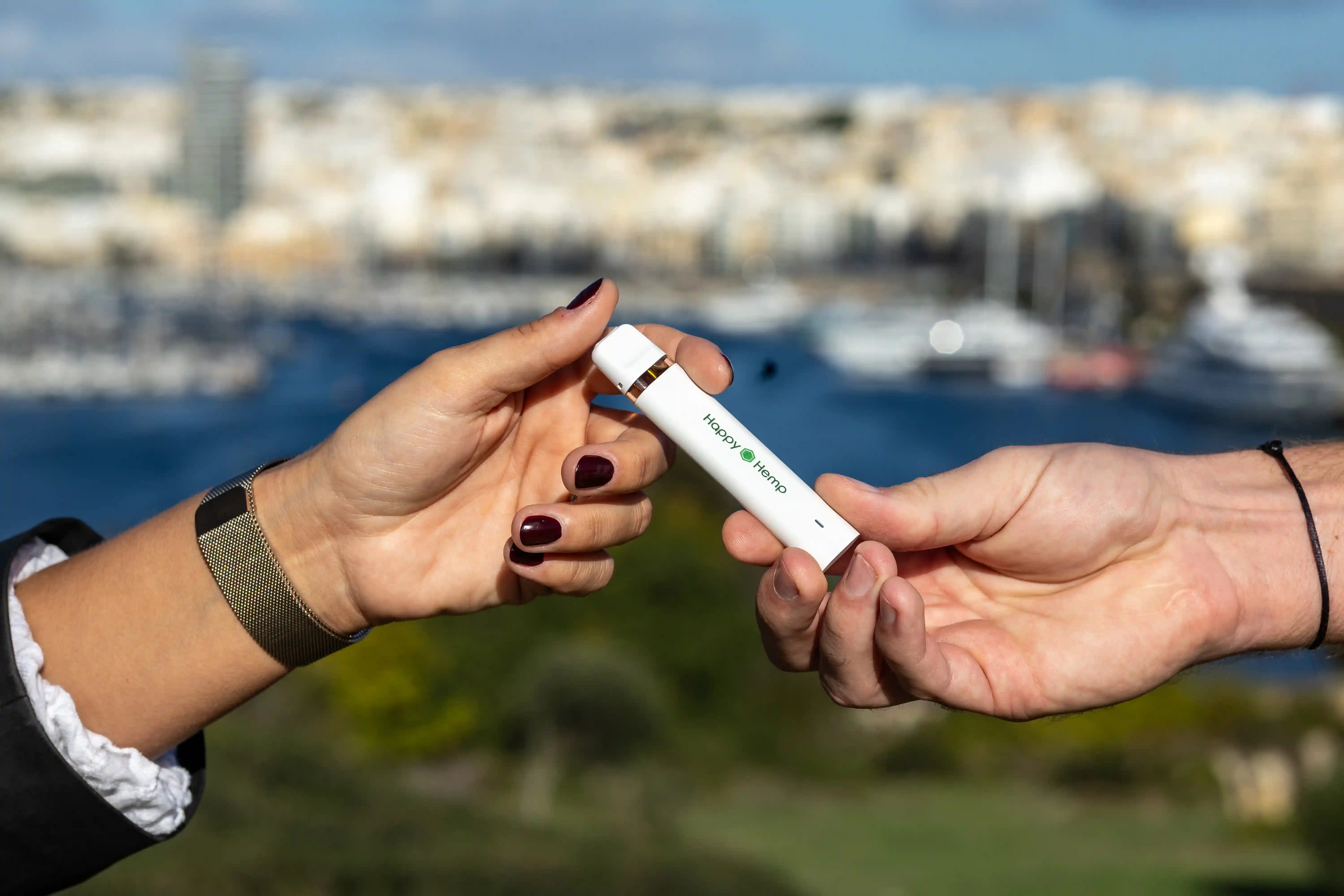Eine weibliche Hand gibt den H3 Ultra Vape Pen in eine männliche Hand. Im Hintergrund ist ein Bootshafen und eine Stadt zu sehen